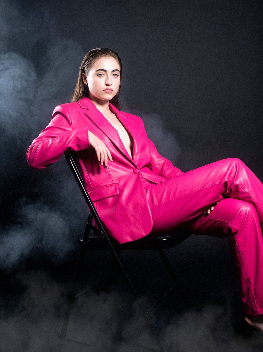 Victoria Grace seated in a tailored pink leather suit, black fog at the floor