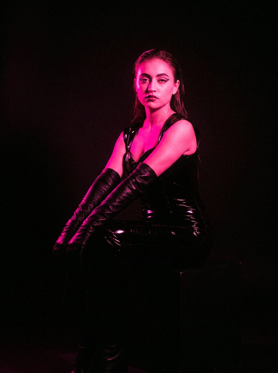 Victoria Grace seated in latex and long leather gloves, lit from the side in pink